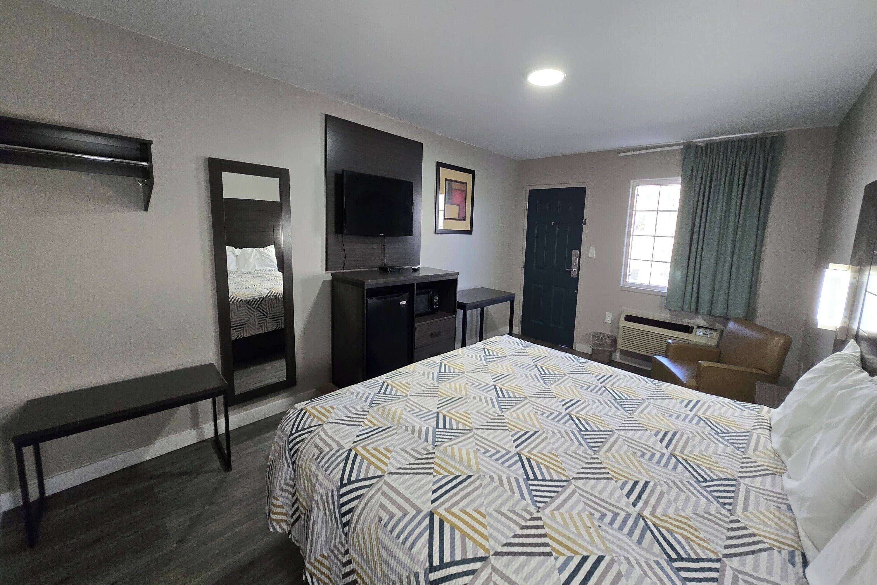 Habitación de hotel con cama, escritorio, silla, televisor, espejo y ventana con cortinas.