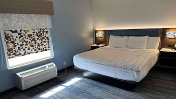 A modern hotel room with a large bed, a heater, a window with geometric blinds, and a nightstand with a lamp.