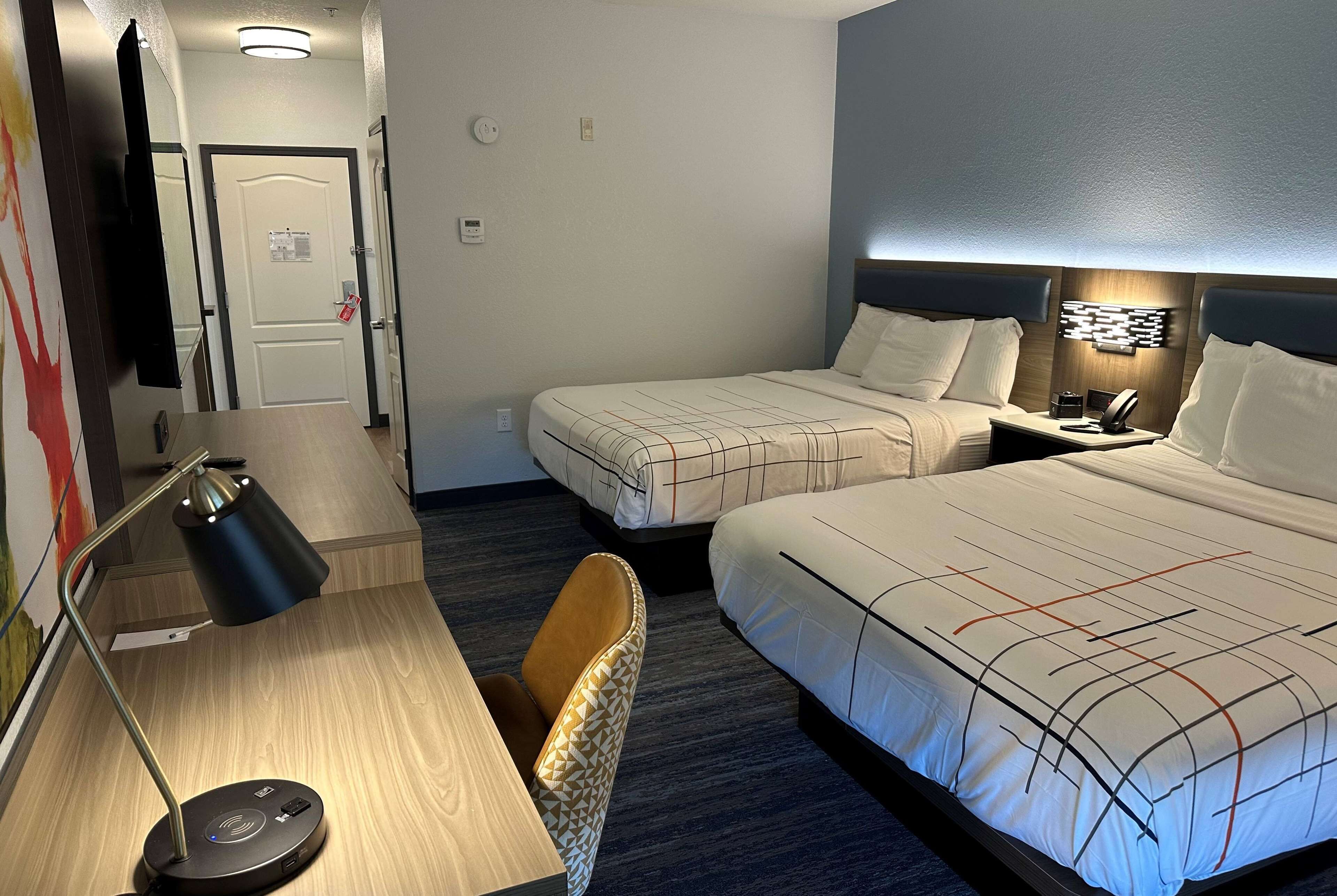 A hotel room with two beds, a desk with a lamp, a chair, and a nightstand with a phone.