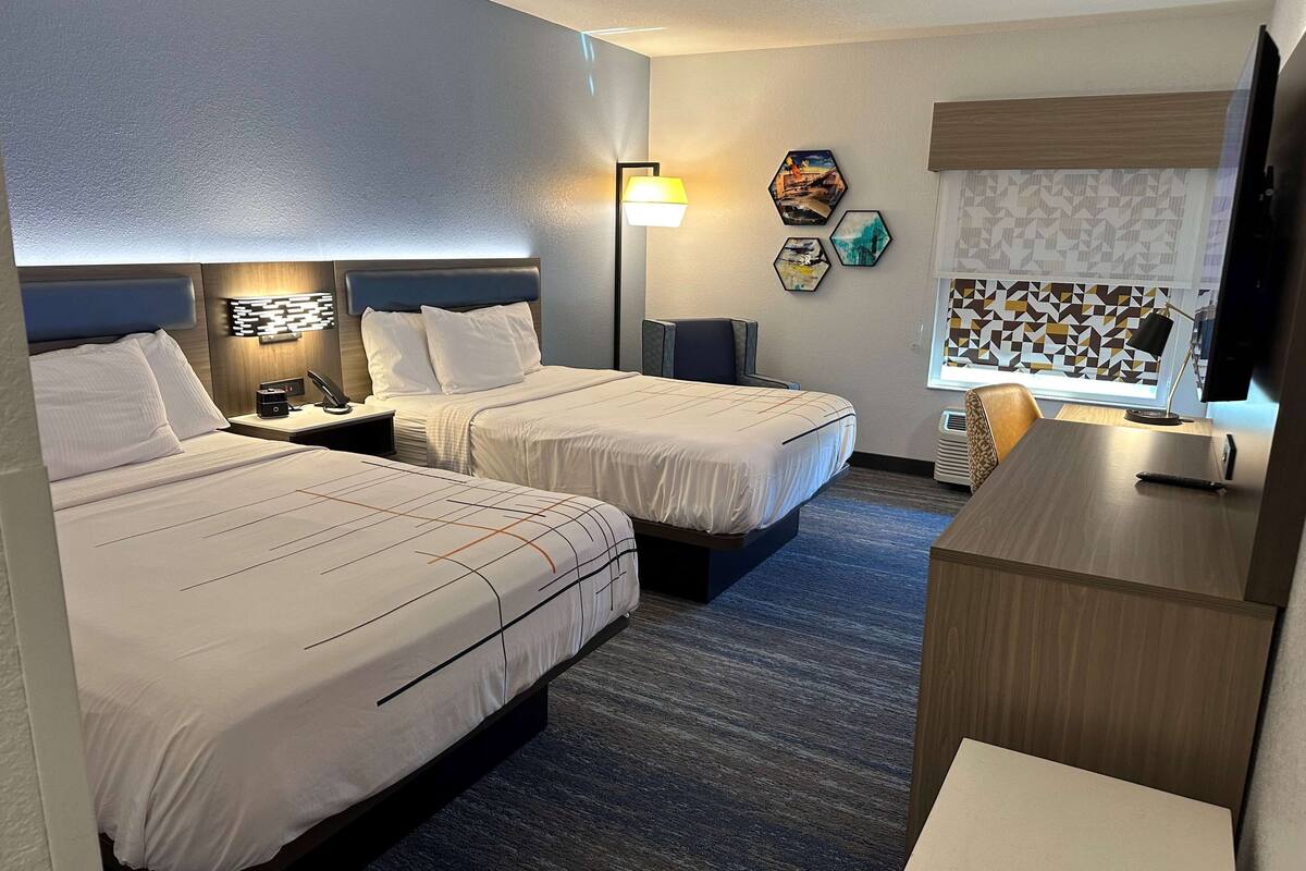 Ein Hotelzimmer mit zwei Betten, einem Schreibtisch, einem Stuhl, einem Fernseher und einem Fenster mit gemusterten Vorhängen.