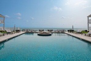 A rectangular swimming pool with a central hot tub, surrounded by lounge chairs and tables, with a view of the sea and a clear sky. - Pelham House Resort (Dennis Port)