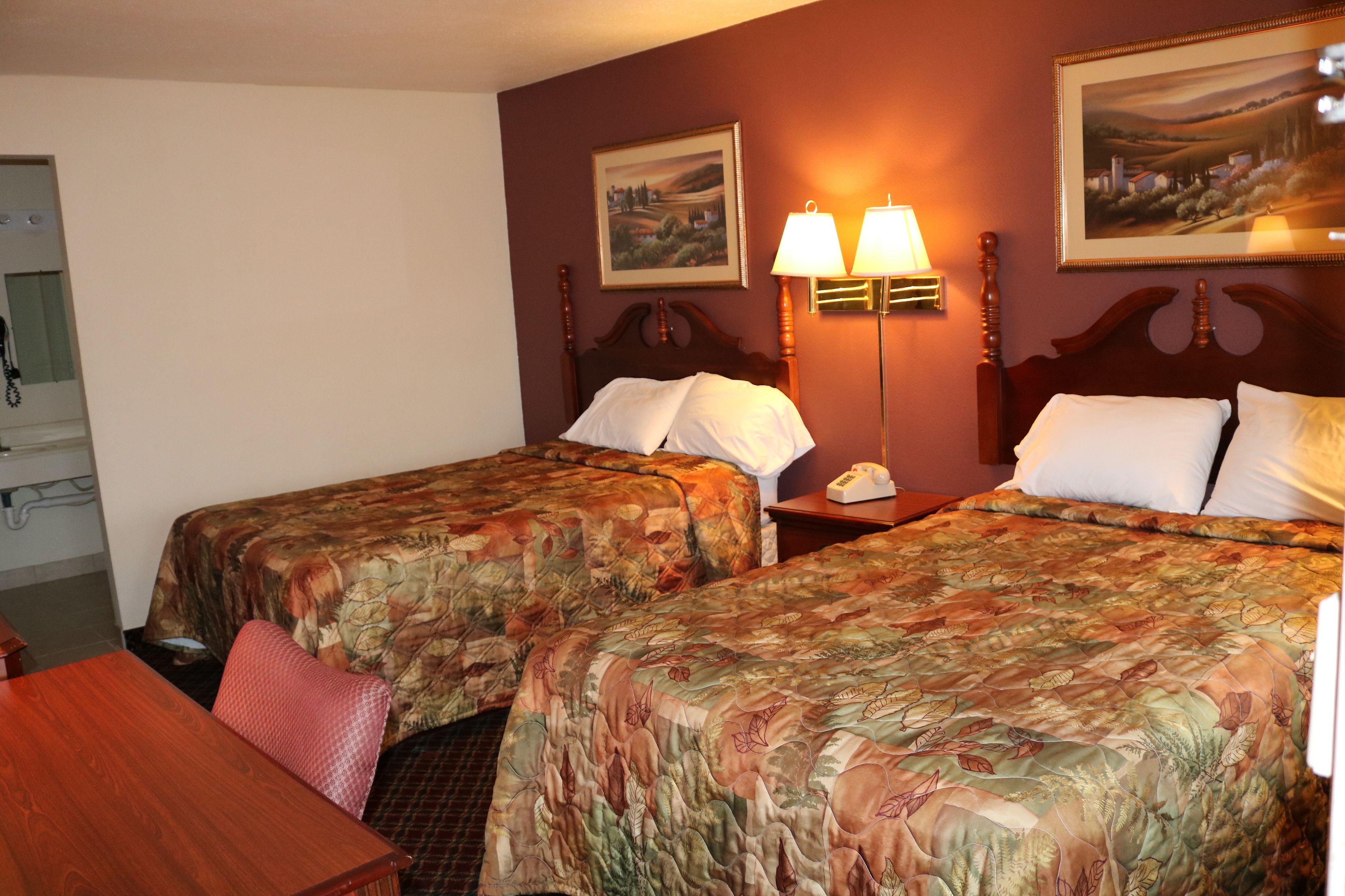 Quarto de hotel com duas camas, uma escrivaninha e uma cadeira. Há duas luminárias fixadas na parede e um quadro sobre as camas.