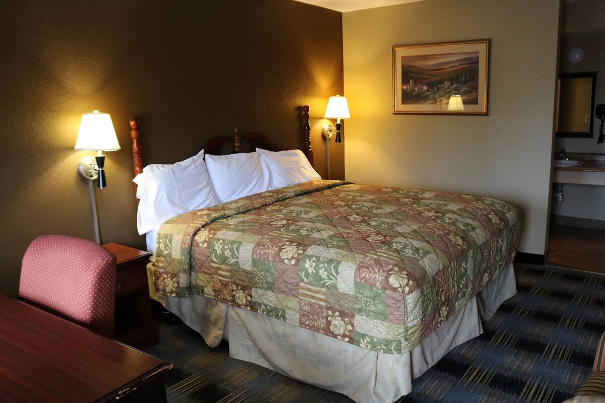 A hotel room with a large bed, two bedside lamps, a wooden headboard, a desk with a chair, and a framed picture on the wall.