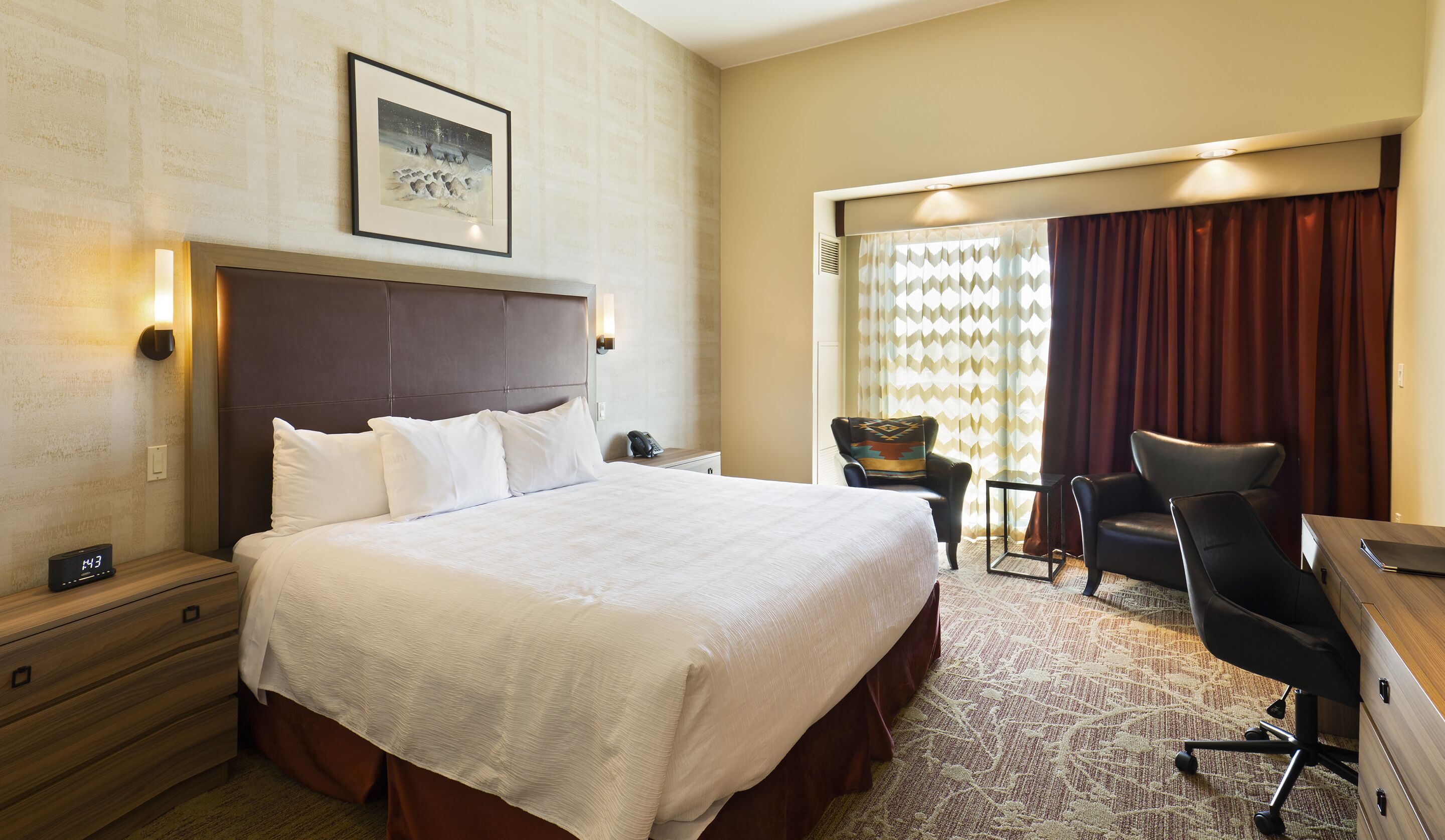 A hotel room with a large bed, a desk, a chair, a nightstand, and a wall-mounted clock.