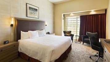 A hotel room with a large bed, a desk, a chair, a nightstand, and a wall-mounted clock.