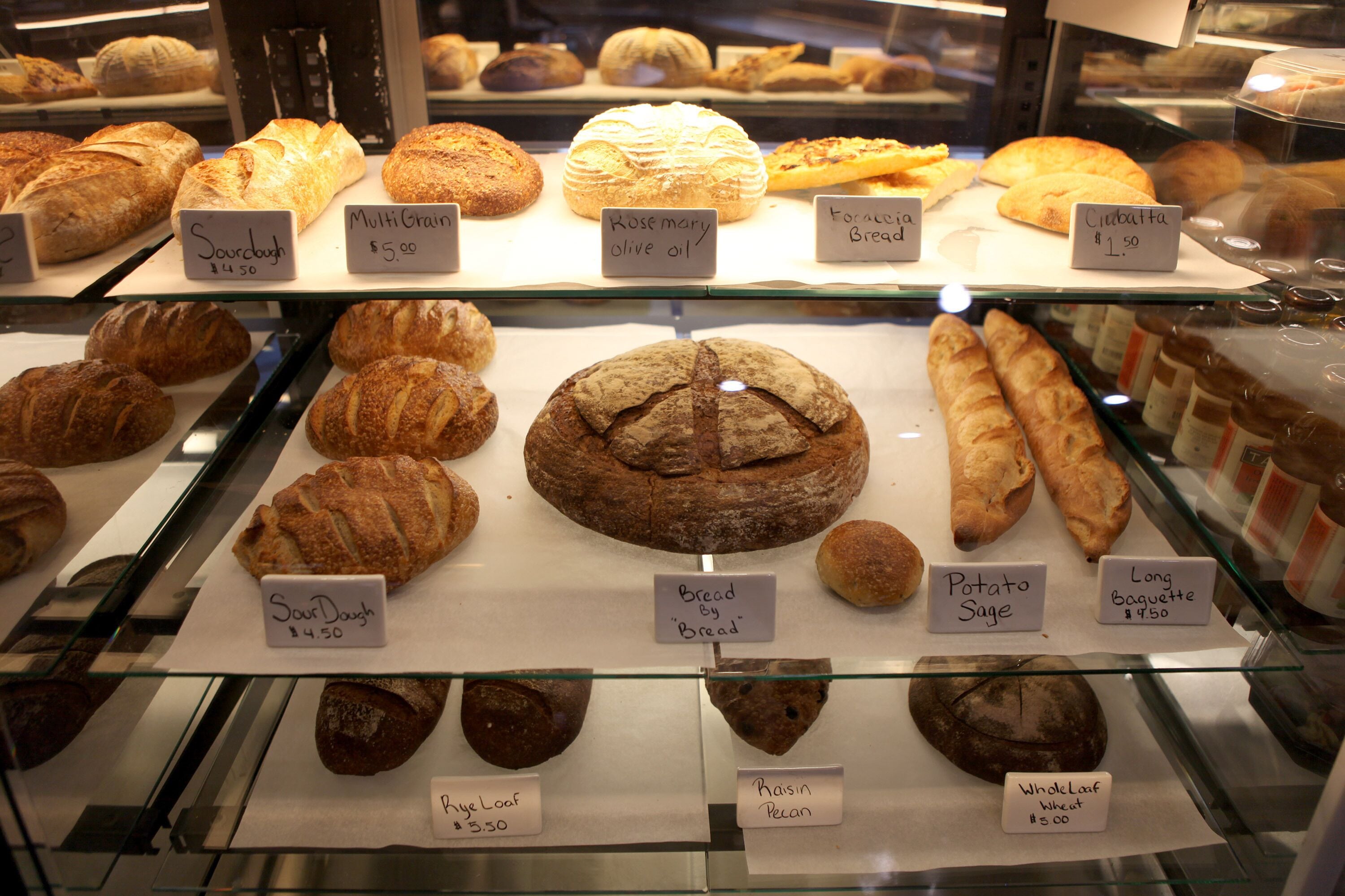 A bakery display case with various types of bread and pastries, each labeled with its name and price.