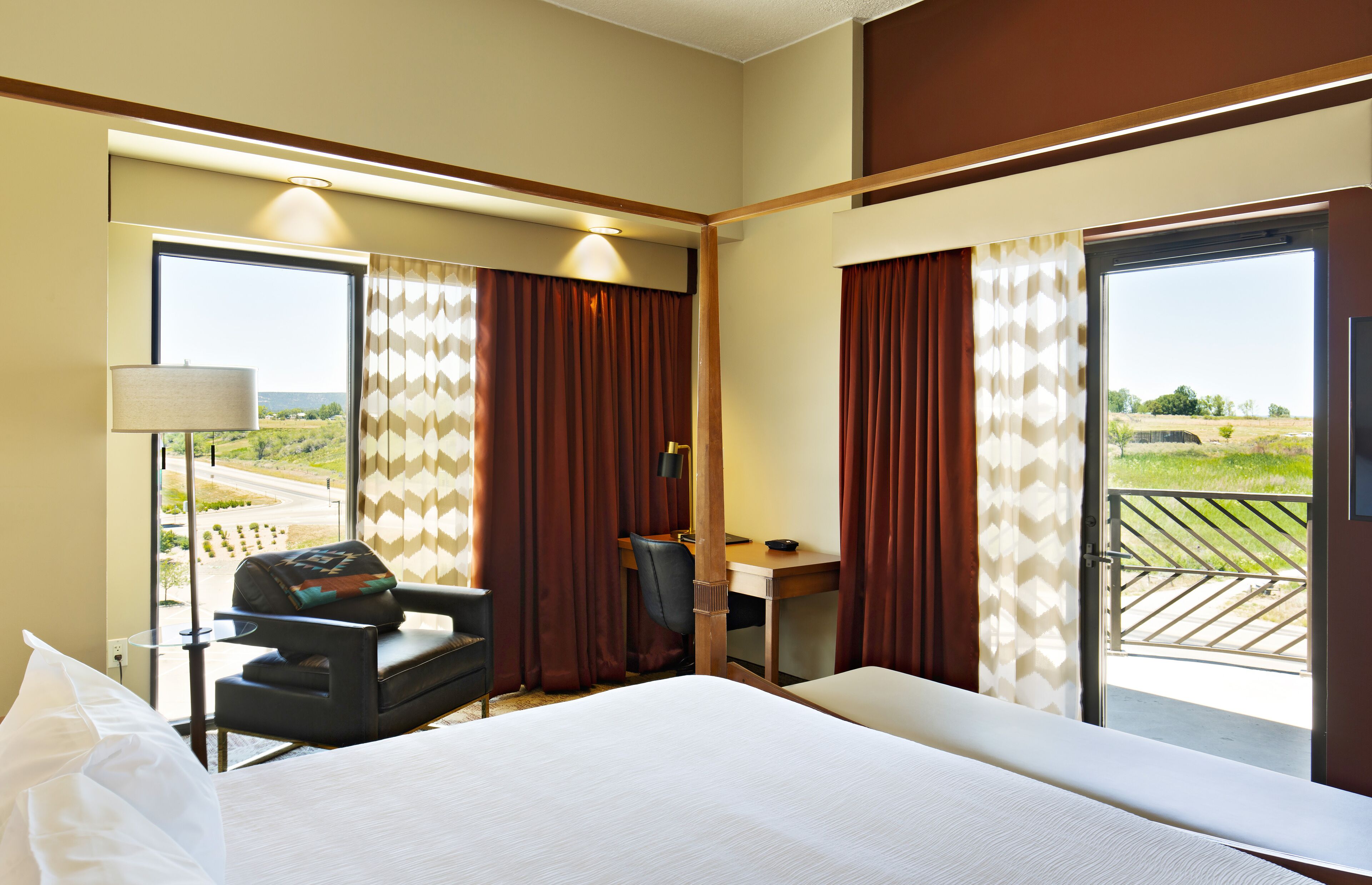 A hotel room with a bed, desk, chair, and large windows offering a view of the outdoors.