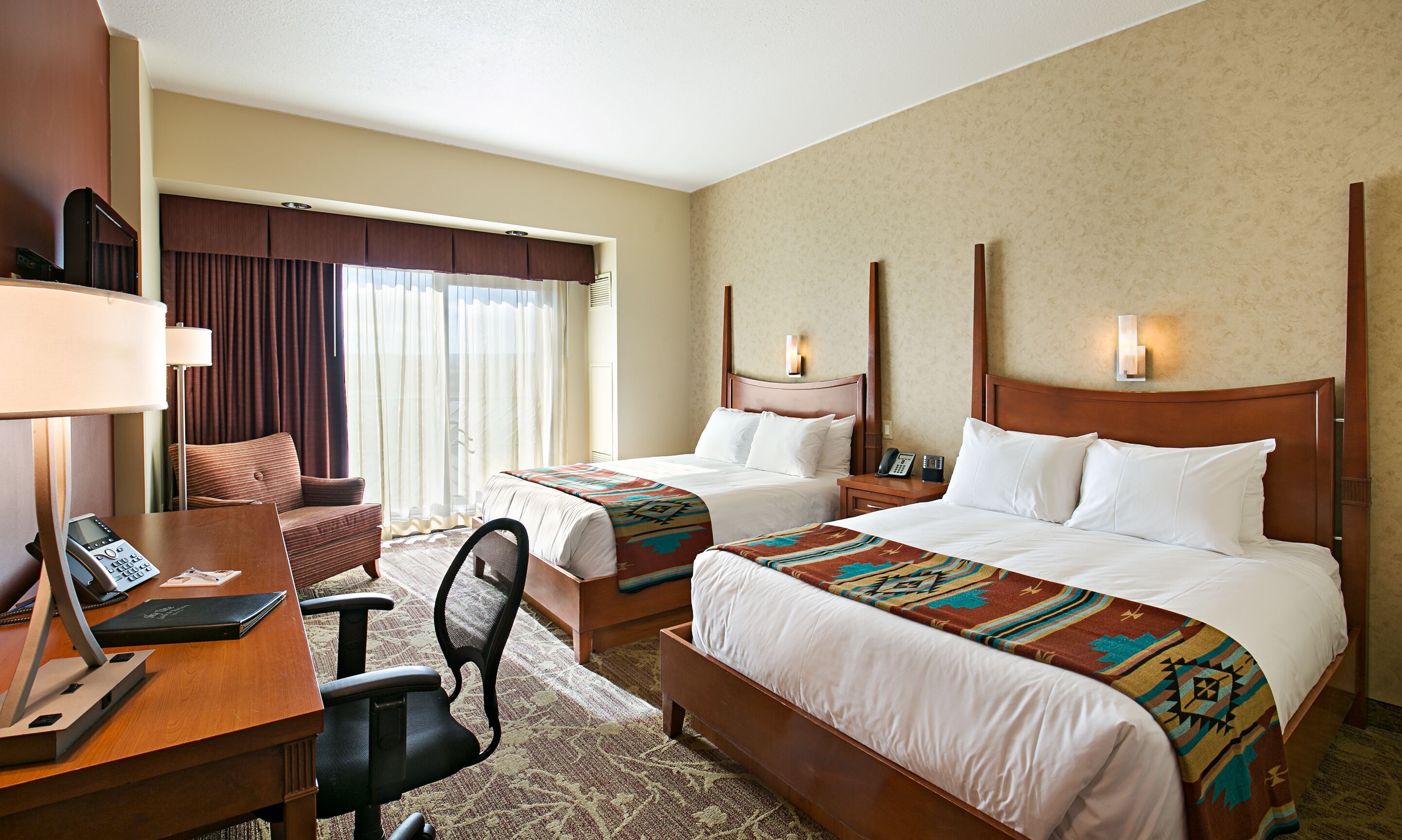 A hotel room with two beds, a desk with a telephone, a chair, and a large window with curtains.