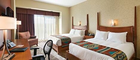 A hotel room with two beds, a desk with a telephone, a chair, and a large window with curtains.