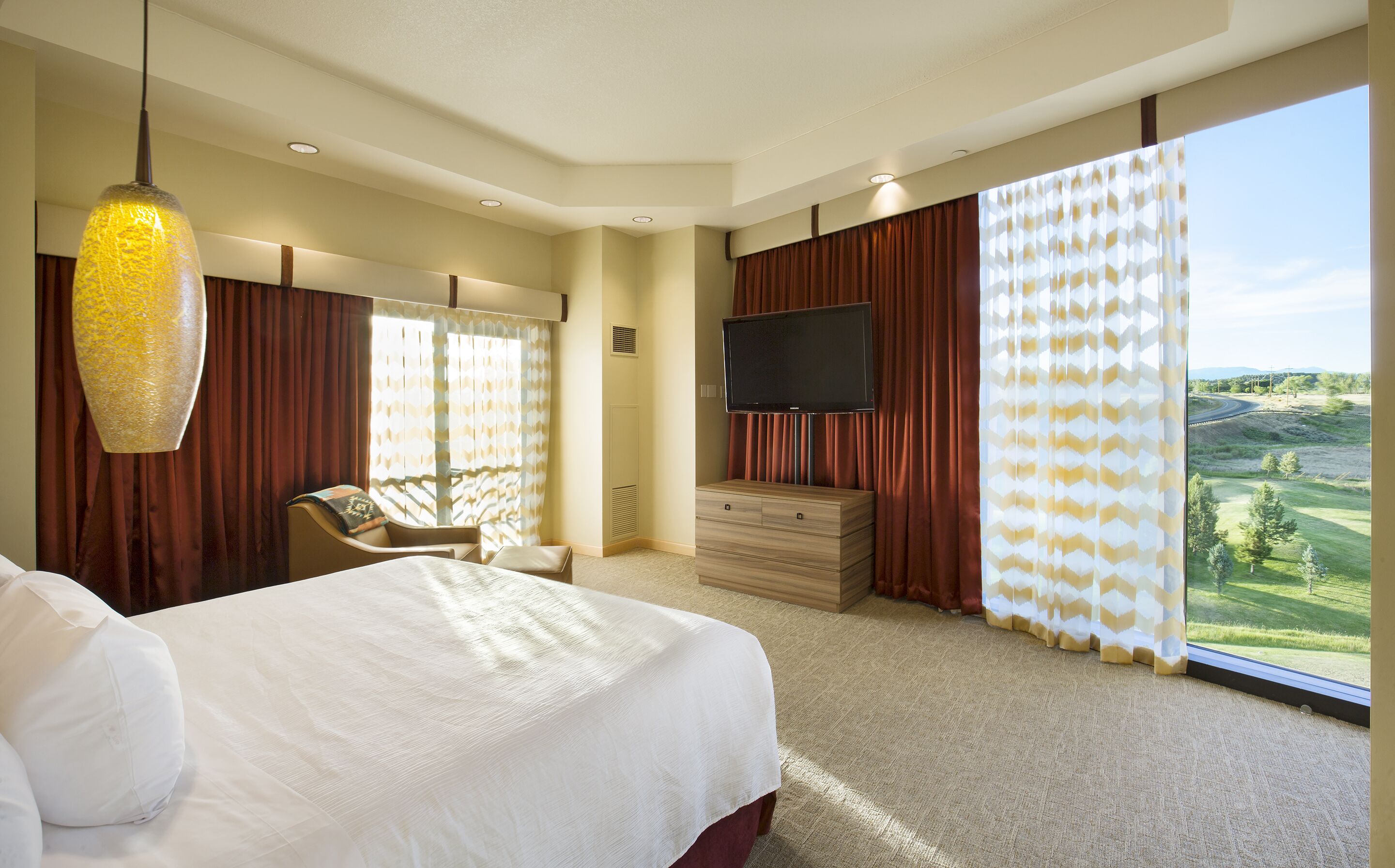 A hotel room with a large bed, a TV, a desk, and a view of a green landscape.