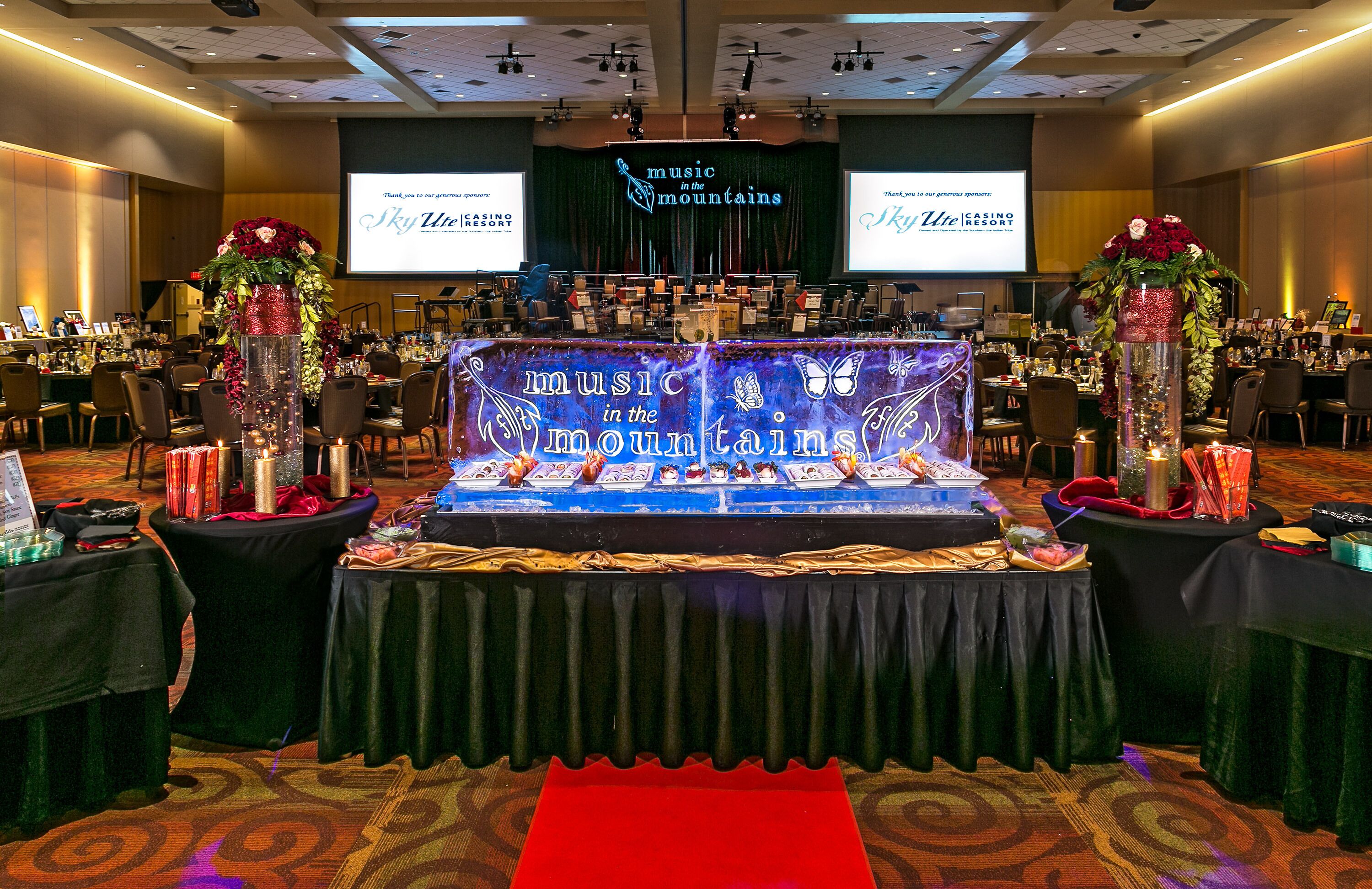 A conference room with a stage at the front, decorated with floral arrangements and a banner reading "Music in the Mountains".