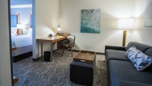 Habitación de hotel con cama, escritorio, silla, sofá, otoman y una obra de arte en la pared.
