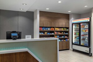 Een hotel receptie met een zelfbedieningskiosk, een koelkast vol snacks en een bord met de naam van het hotel.