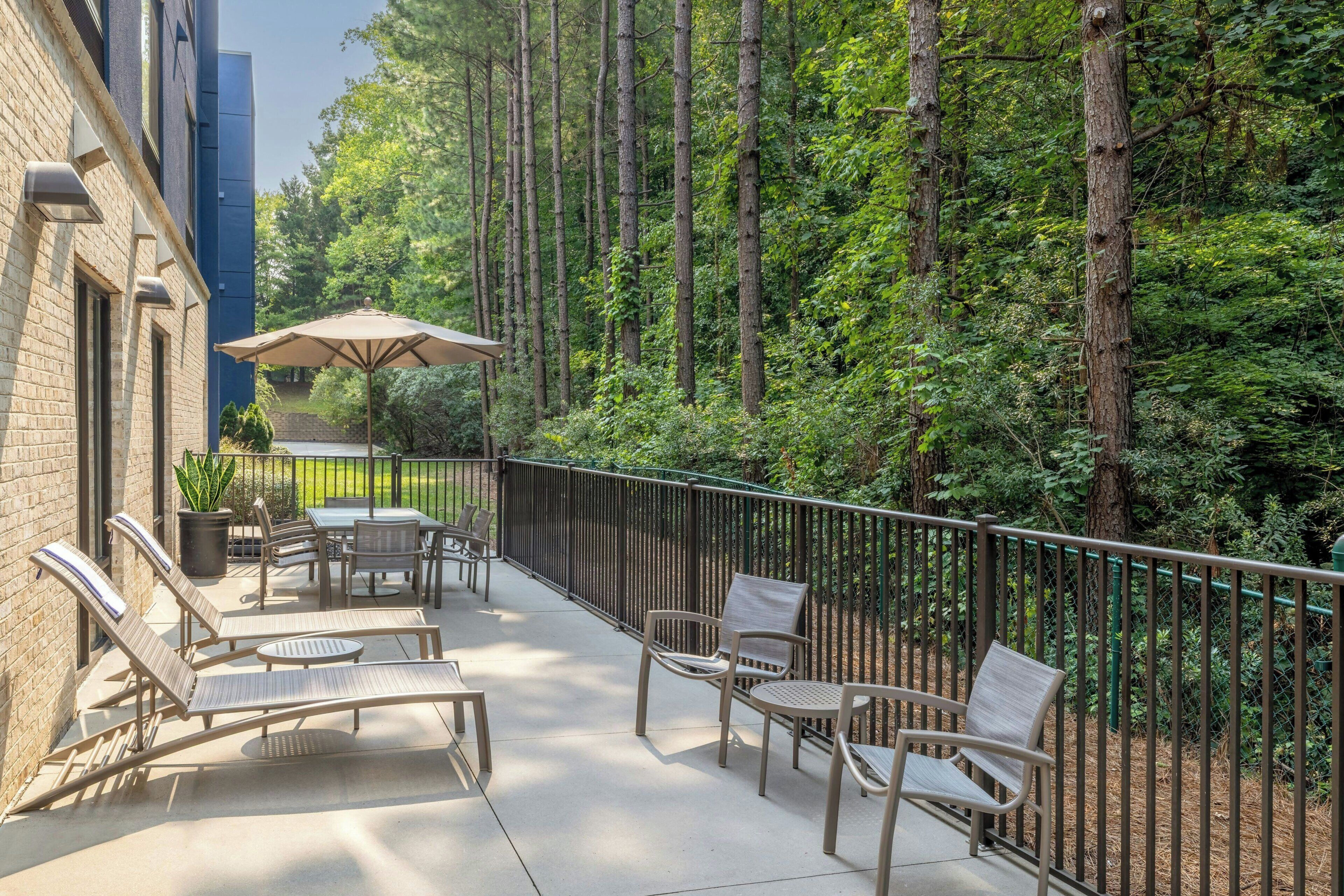 A patio with outdoor furniture, a table, and an umbrella, surrounded by a metal fence and trees.