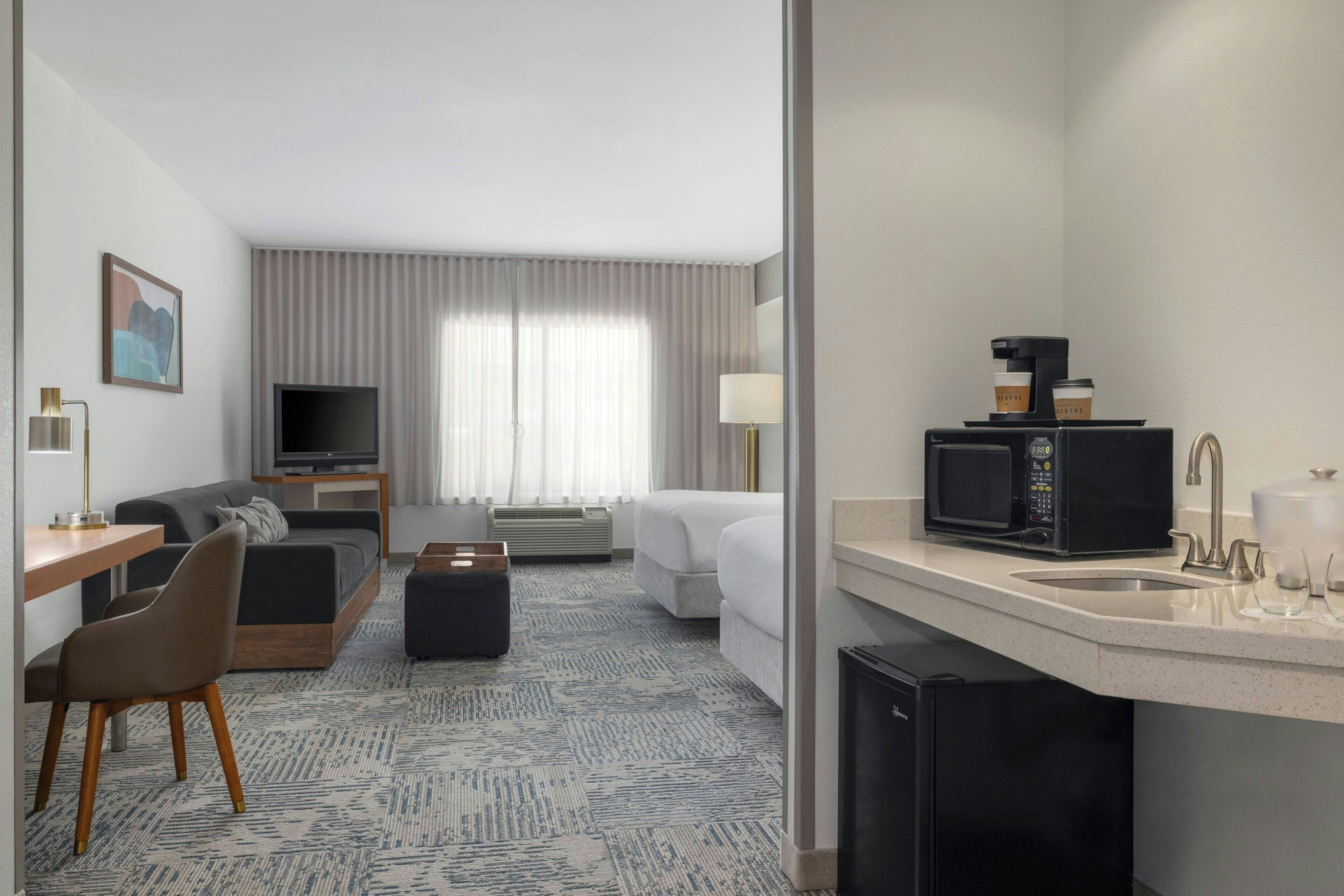 Habitación de hotel con sofá, escritorio, televisor, cafetera y kitchenette.