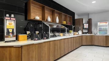 Un área de desayuno autoservicio con dispensadores de gofres, una cafetera y un refrigerador.