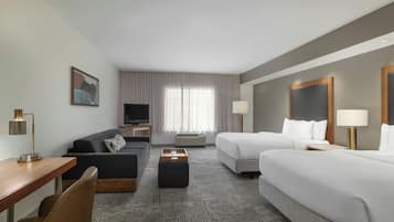 Hotelværelse med sofa, to senge, et skrivebord, et fjernsyn og et vindue med gardiner.