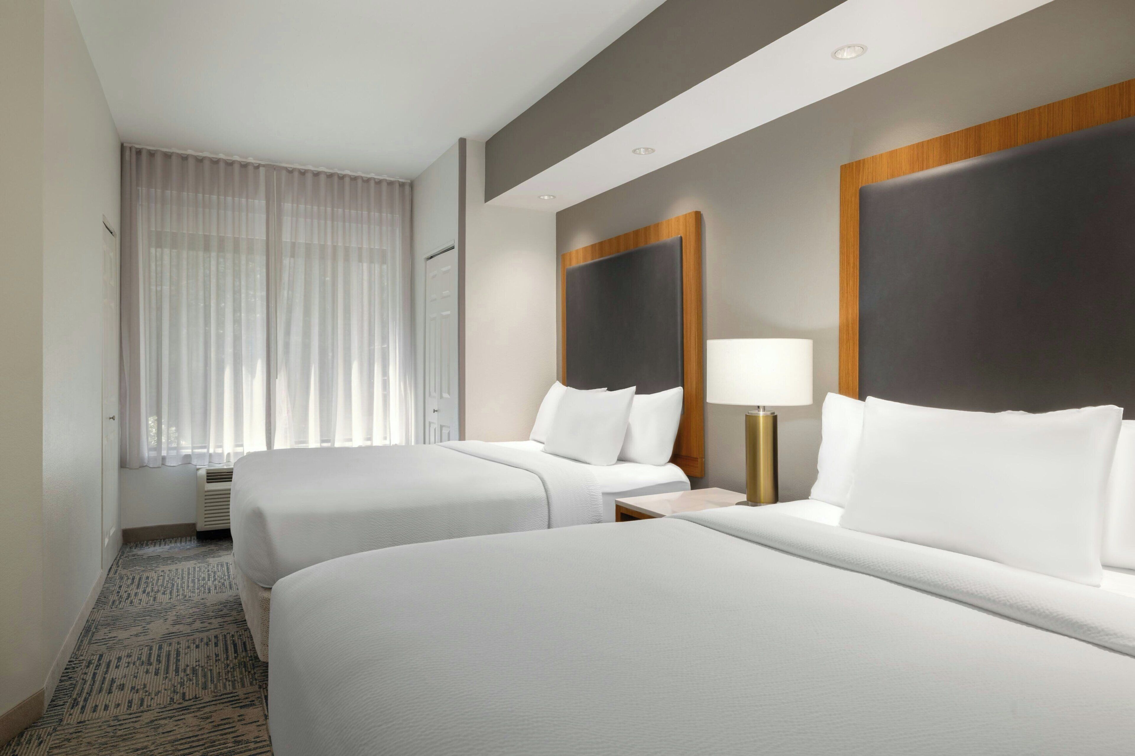 Habitación de hotel con dos camas, un ventanal con cortinas traslúcidas, una mesita de noche con lámpara y piso alfombrado.