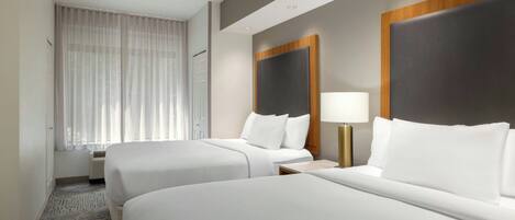 Habitación de hotel con dos camas, un ventanal con cortinas traslúcidas, una mesita de noche con lámpara y piso alfombrado.