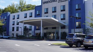 Un hotel moderno con vestíbulo cubierto, varios pisos y un estacionamiento con vehículos utilitarios deportivos.