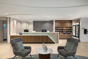 A hotel reception area with a curved counter, two armchairs, a coffee table, and a minibar stocked with snacks. - Springhill Suites by Marriott Winston-Salem Hanes Mall (Winston-Salem)