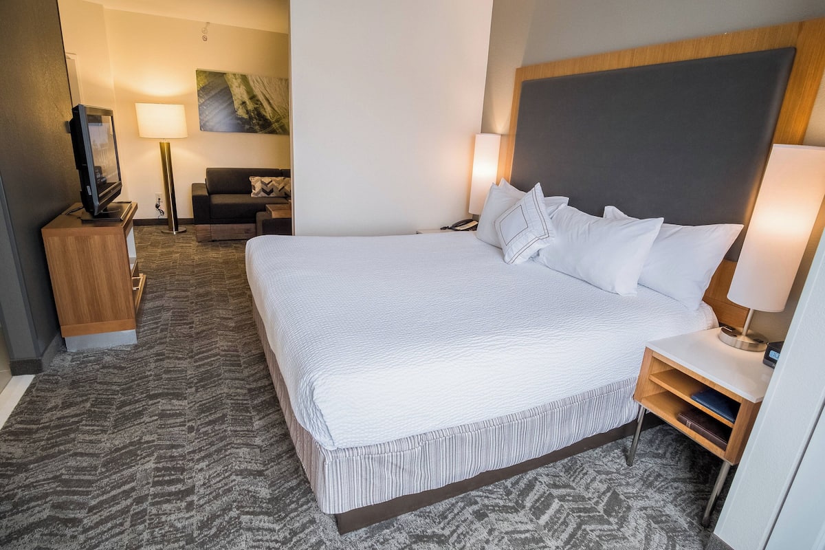 Ein Hotelzimmer mit einem großen Bett, einem Fernseher, einem Schreibtisch mit Lampe und einer Couch im Hintergrund.
