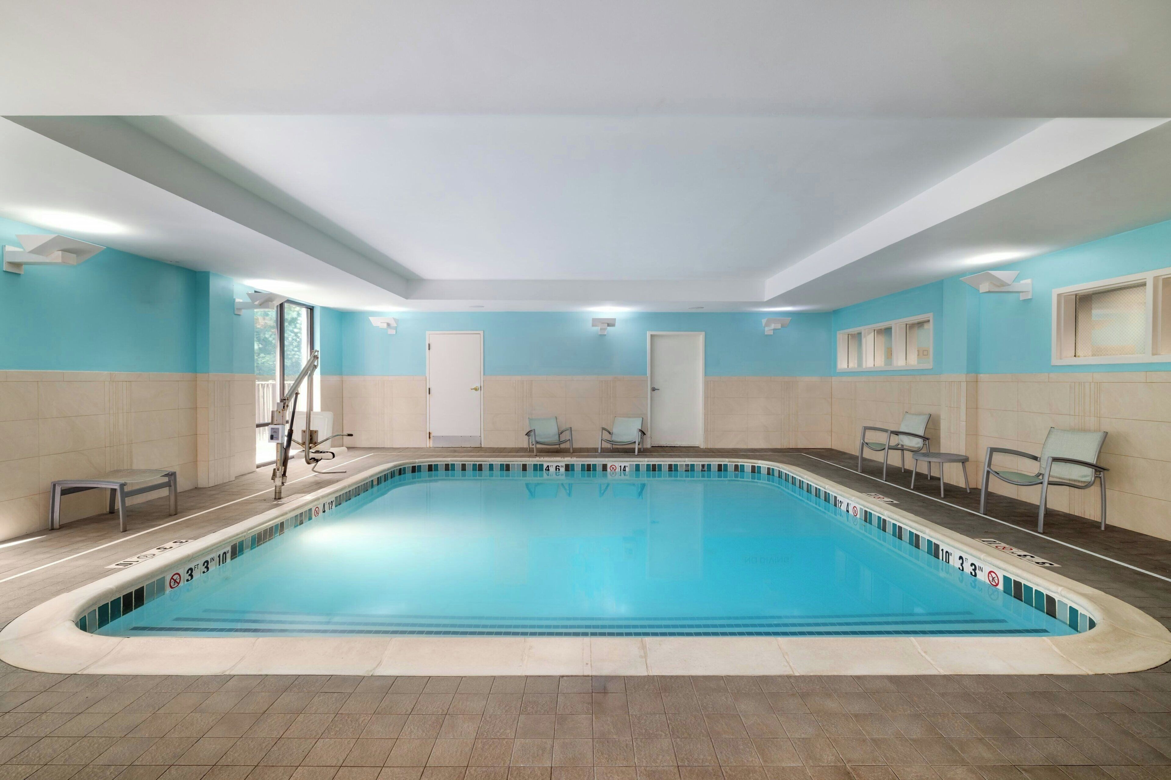 En stor indendørs swimmingpool med krystalklart vand, omgivet af beige og lyseblå vægge, og med et springbræt samt liggestole.