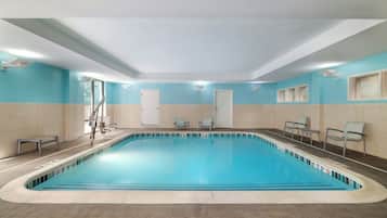 En stor indendørs swimmingpool med krystalklart vand, omgivet af beige og lyseblå vægge, og med et springbræt samt liggestole.