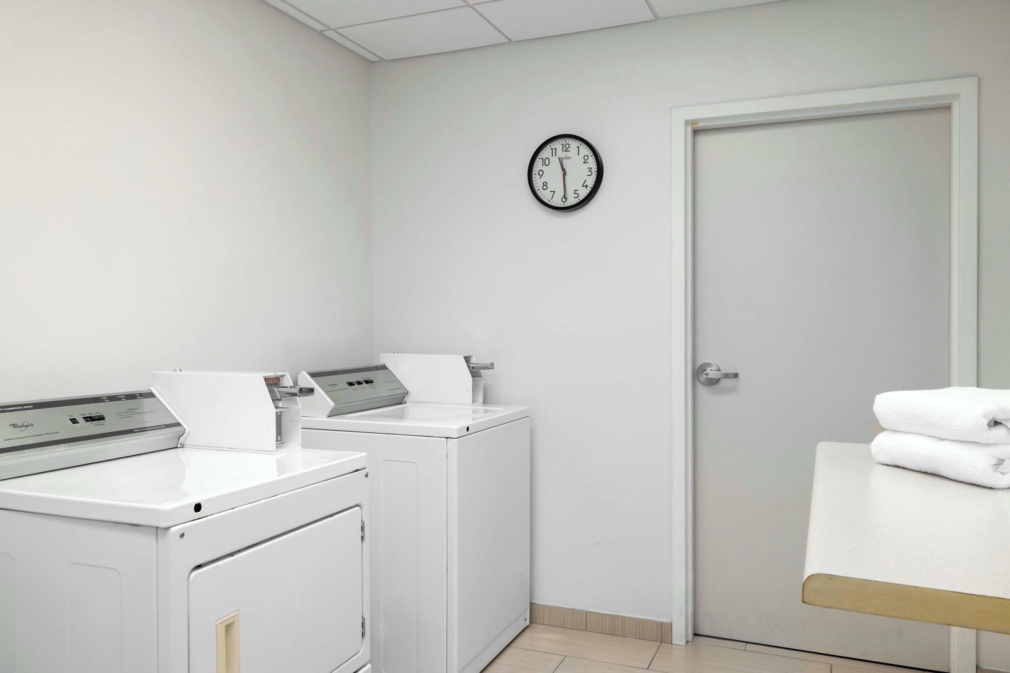 A laundry room with two washing machines, a dryer, a clock on the wall, and a door.