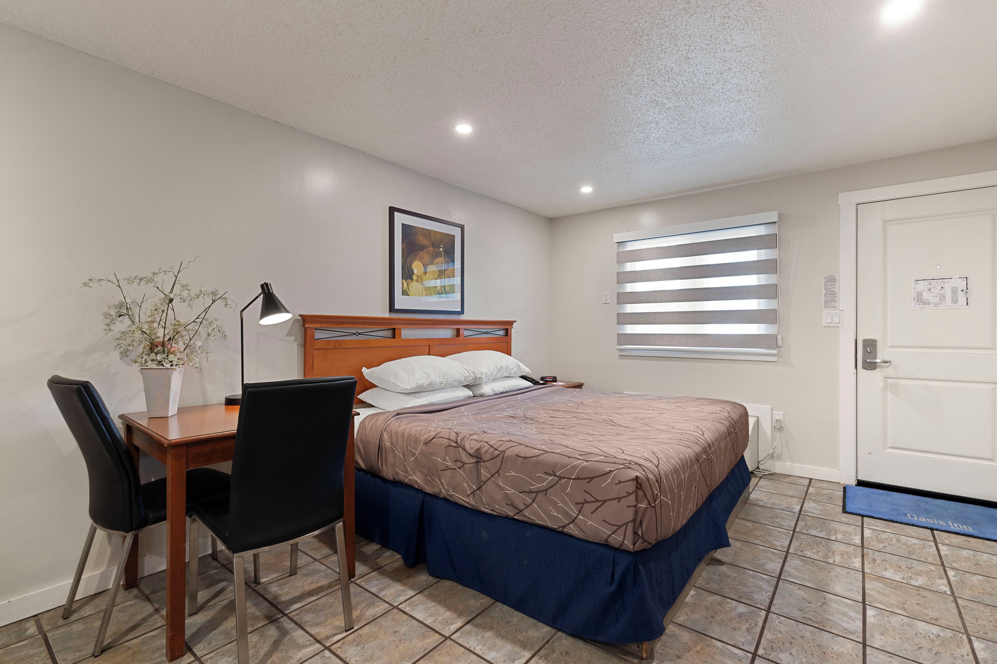 Un dormitorio con una cama, un escritorio y una silla. Hay una ventana con persianas, una puerta y una planta sobre el escritorio.
