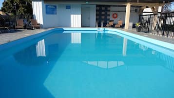 Una piscina con superficie de agua azul cristalina, rodeada de sillas de descanso y un edificio con un cartel que indica las normas de la piscina.