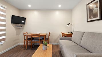 Ein Wohnzimmer mit grauem Sofa, einem Holzesstisch mit Stühlen, einem flachbildfernseher an der Wand und einem gerahmten Bild über dem Sofa.