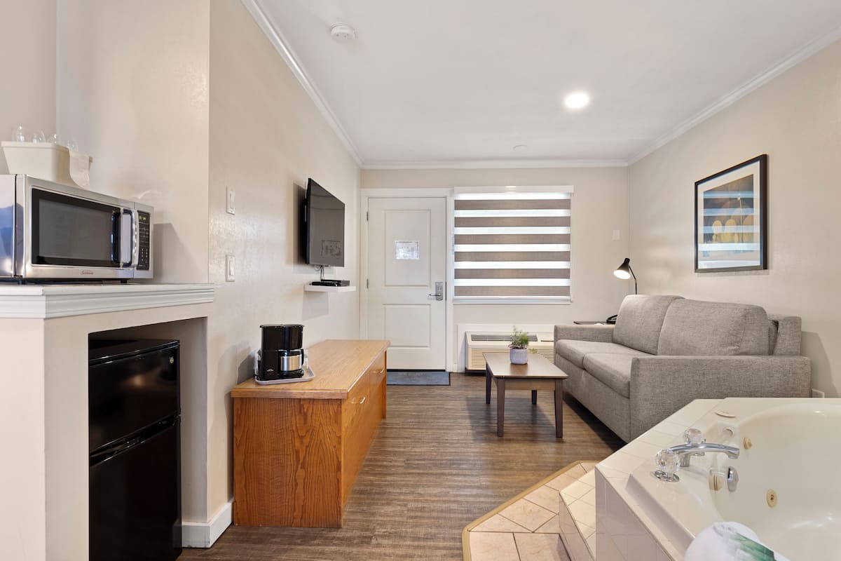 A modern kitchen with a microwave, coffee maker, and a black dishwasher. There is a wooden counter, a gray sofa, and a white bathtub in the living area.