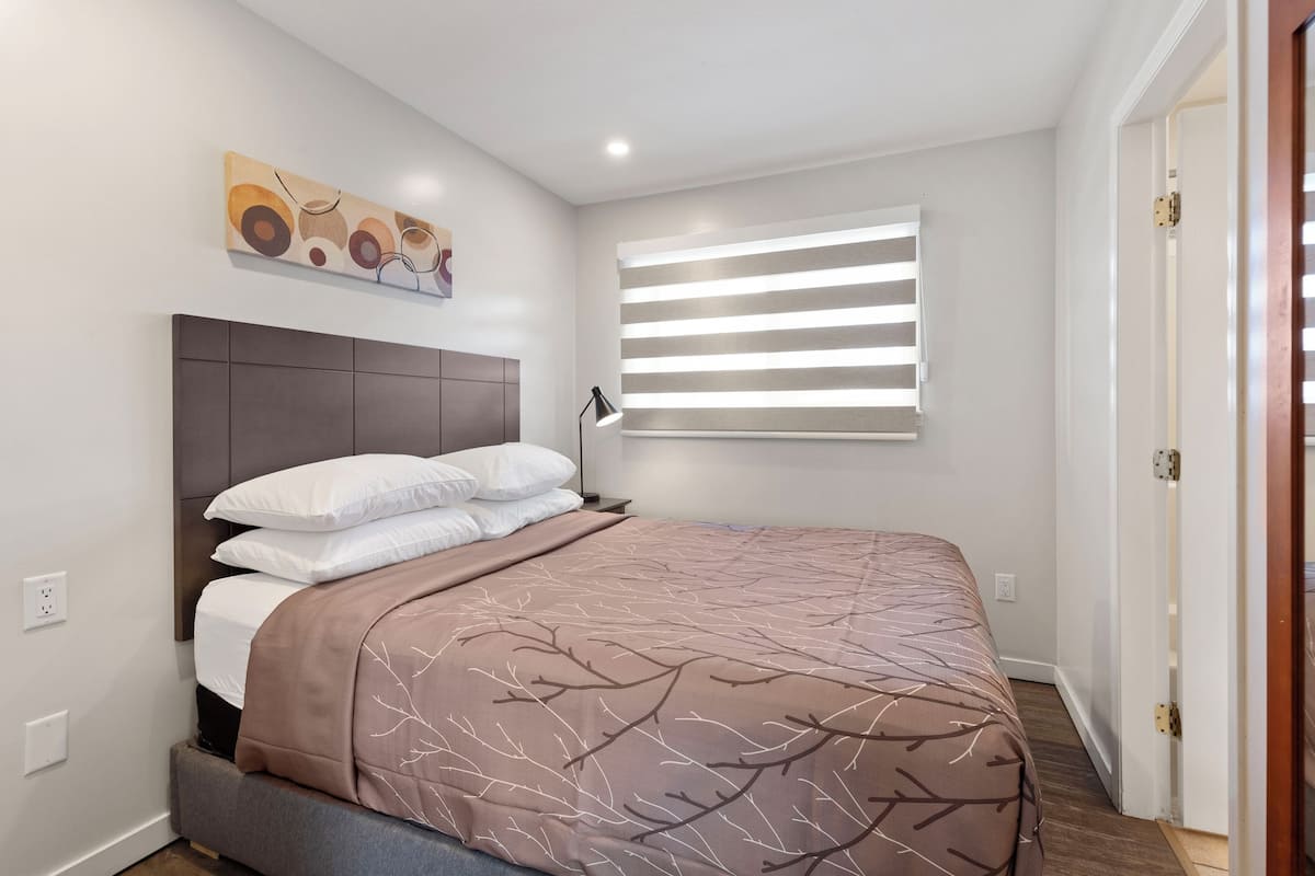 A bedroom with a bed, a headboard, a window with blinds, and a painting on the wall.