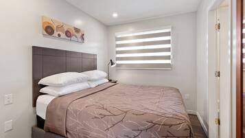A bedroom with a bed, a headboard, a window with blinds, and a painting on the wall.