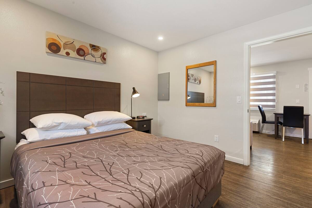A bedroom with a large bed, a bedside table with a lamp, a wall-mounted artwork, and a mirror.
