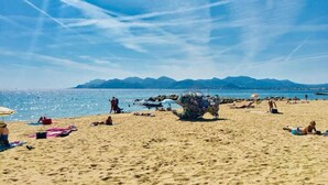 A beach with people sunbathing and swimming, a clear sky, and mountains in the background. - Hôtel Cannes Croisette (Cannes)