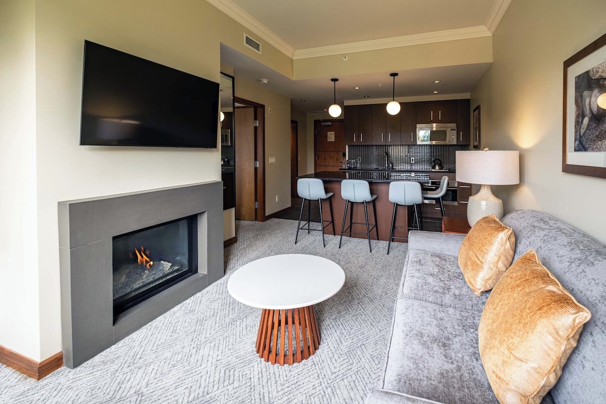 Een moderne woonkamer met een open haard, een flatscreen televisie, een ronde salontafel, een bank met kussens, en een keuken in de achtergrond.
