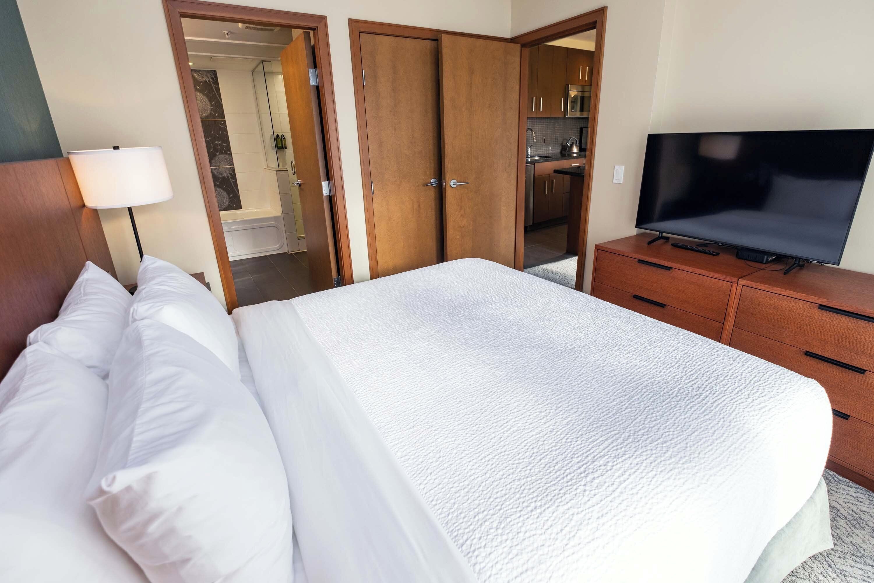 Une chambre d’hôtel avec un lit, un téléviseur posé sur une commode, une salle de bain visible à travers une porte ouverte et un coin cuisine.