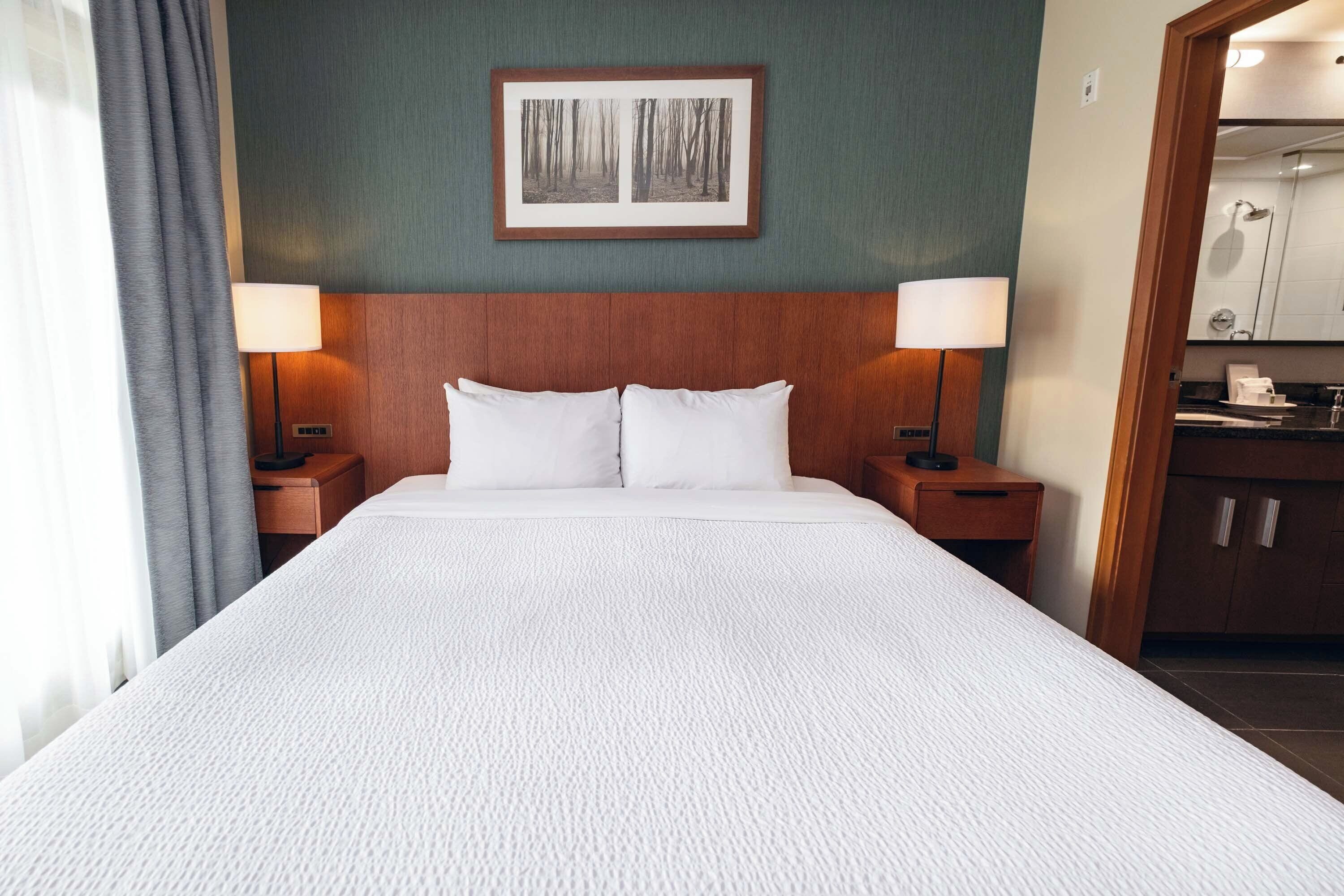 A hotel room with a large bed, two bedside tables with lamps, a framed picture on the wall, and a view into the bathroom.