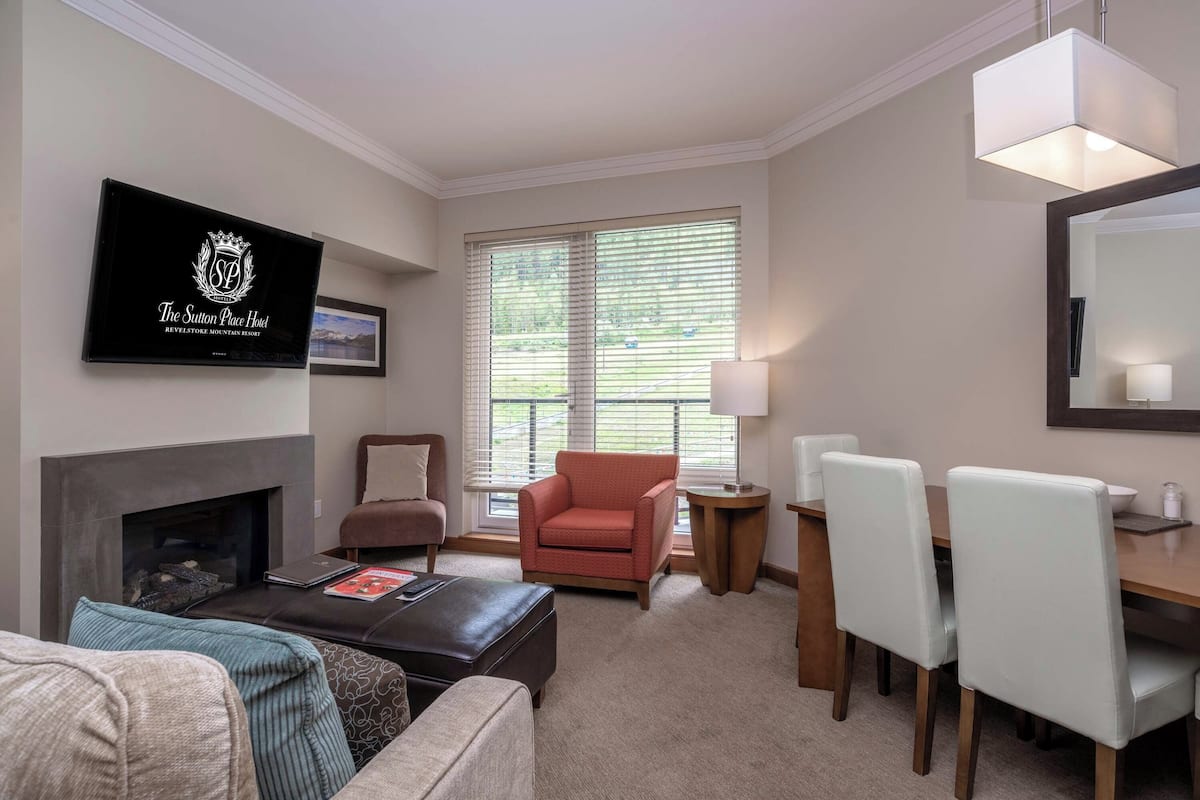 Een hotelkamer met een open haard, een aan de muur bevestigde televisie, een zithoek met tafel en stoelen, en een raam met zonwering.