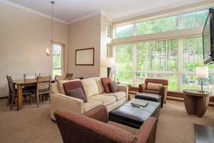 Amplia sala de estar con un sofá color beige, sillones estampados, una mesa de centro y una zona de comedor con mesa y sillas de madera. Ventanales grandes ofrecen vistas a zonas verdes.