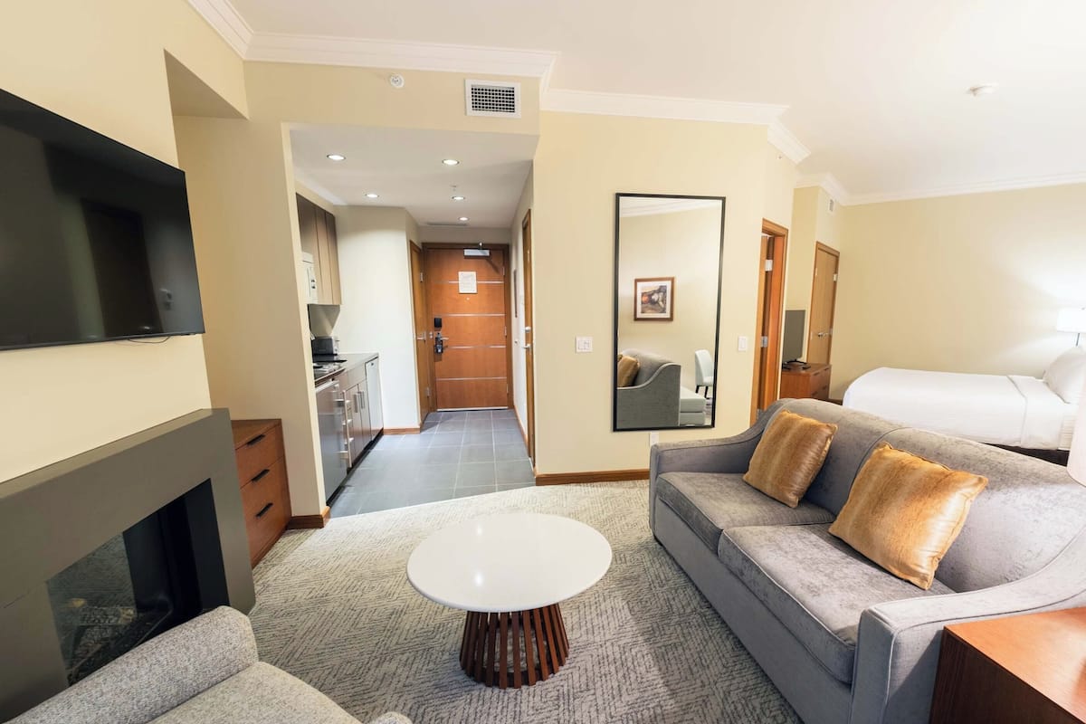 Een moderne hotelkamer met een grijze bank, een witte salontafel, een flatscreen-tv, een open haard en een keukenruimte zichtbaar door een geopende deur.