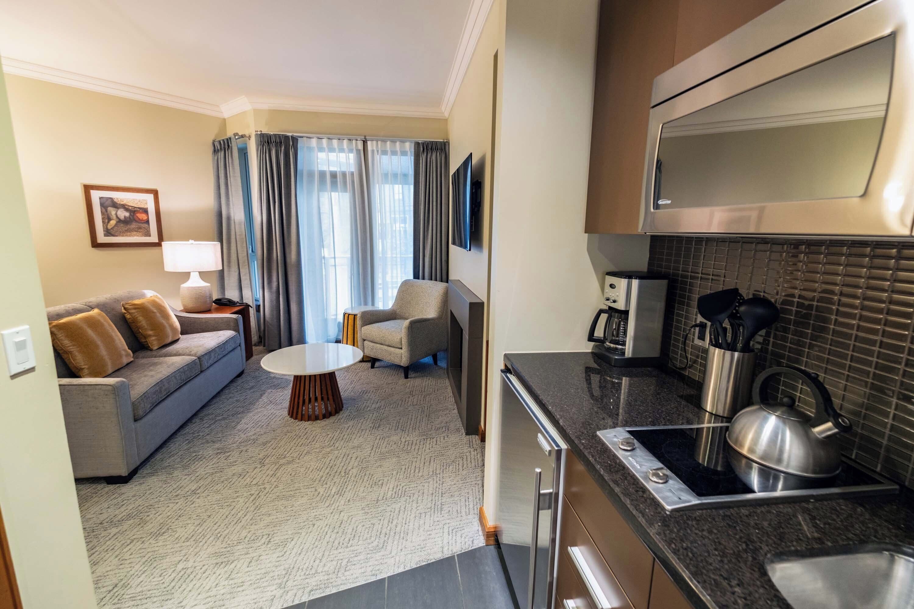 A compact living space with a sofa, armchair, coffee table, kitchenette, and a TV mounted on the wall.