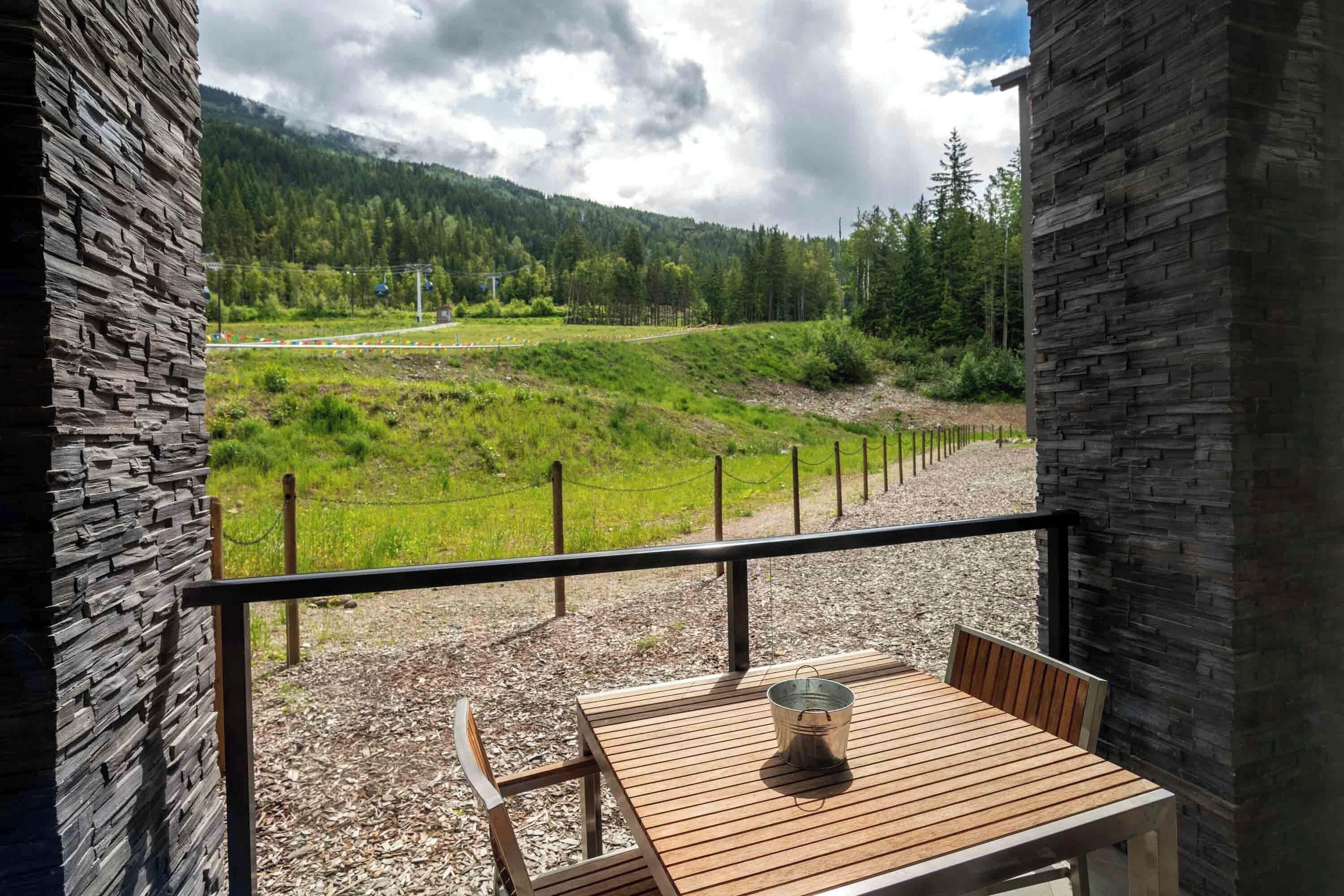 Un balcón con una mesa y sillas de madera, con vistas a un paisaje verde que incluye una cerca y una carretera a lo lejos.