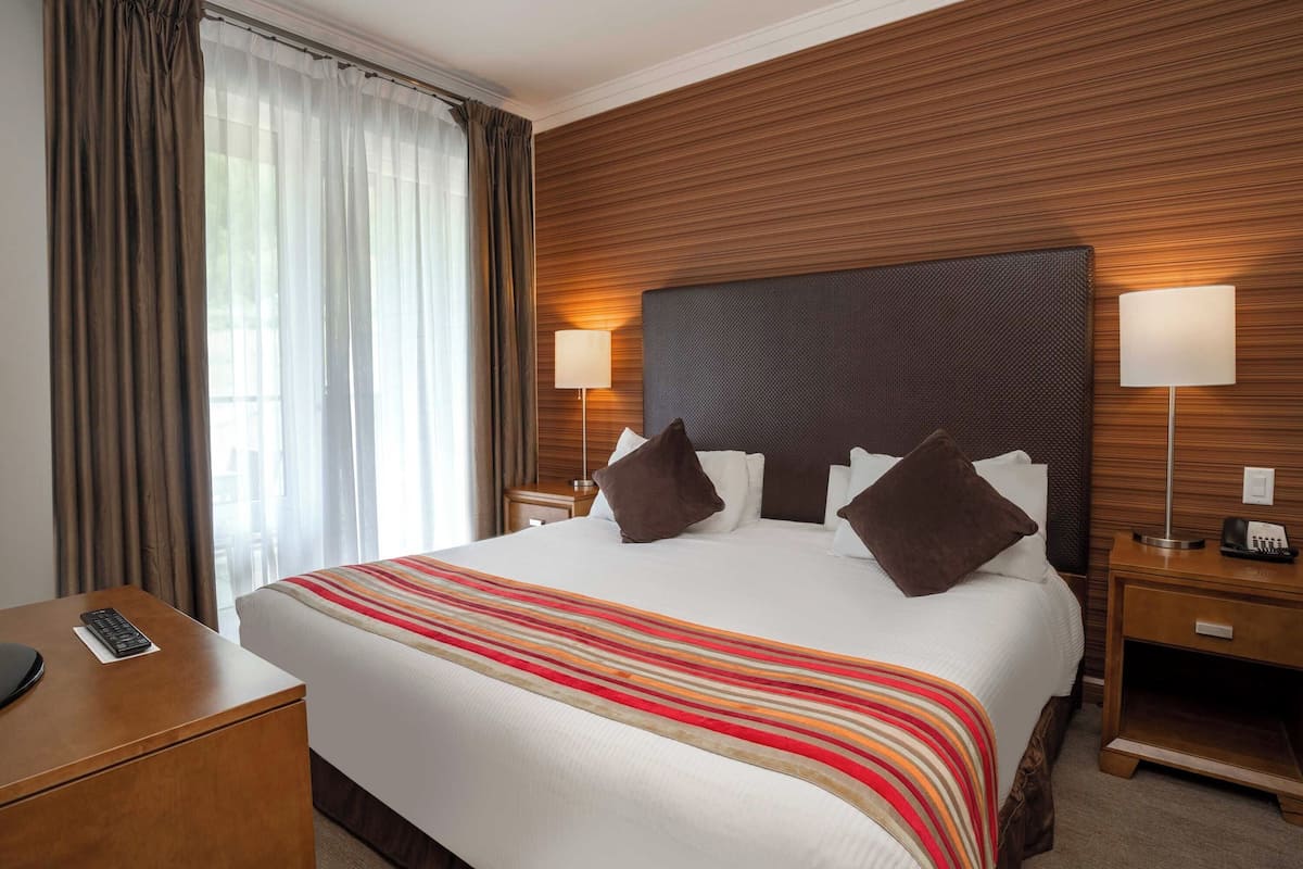 Een hotelkamer met een groot bed, een houten hoofdeinde, twee nachtkastjes met lampen, een bureau met een afstandsbediening en een raam met gordijnen.