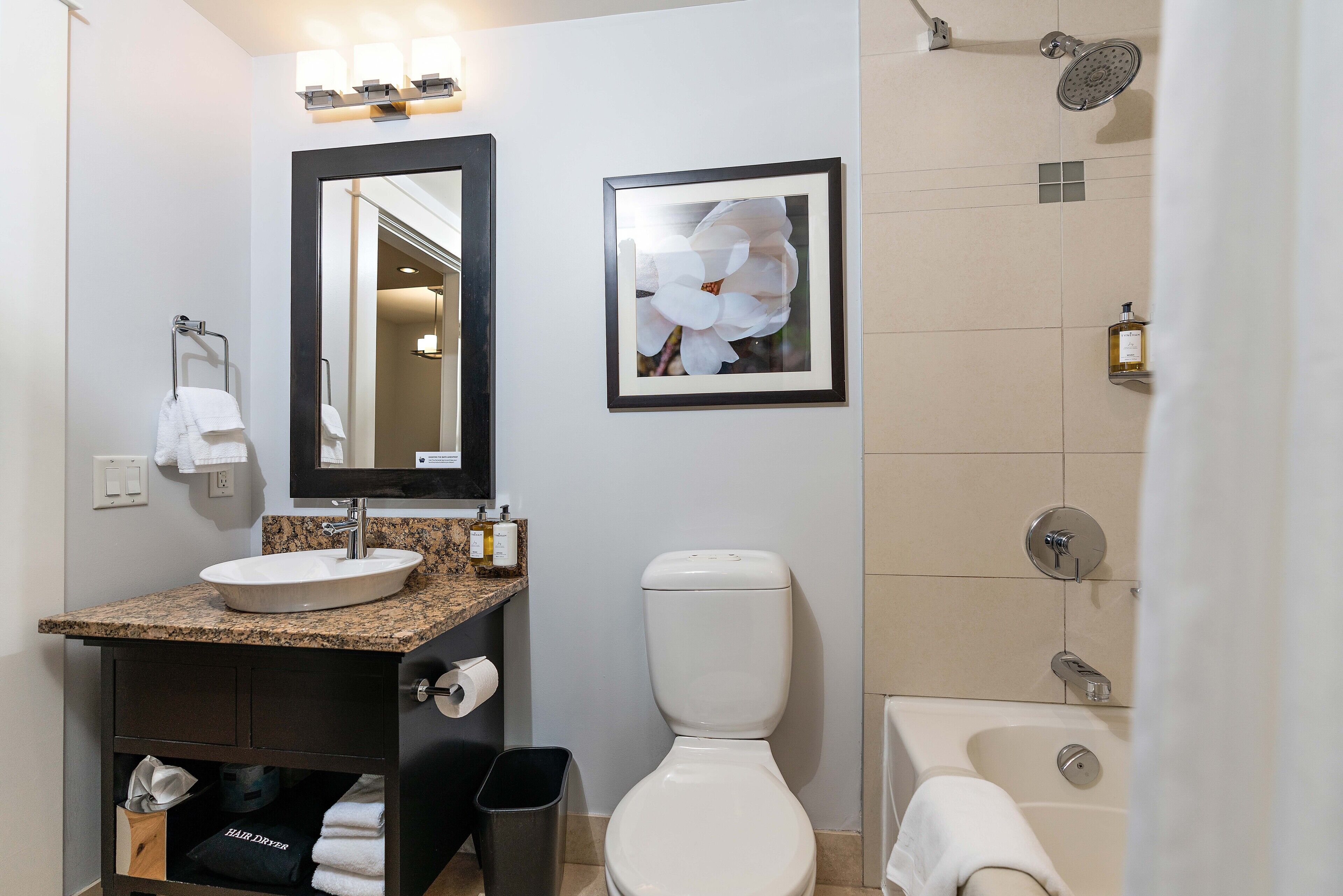 Un baño moderno con lavamanos de granito, inodoro, un cuadro enmarco de una flor y zona de ducha.