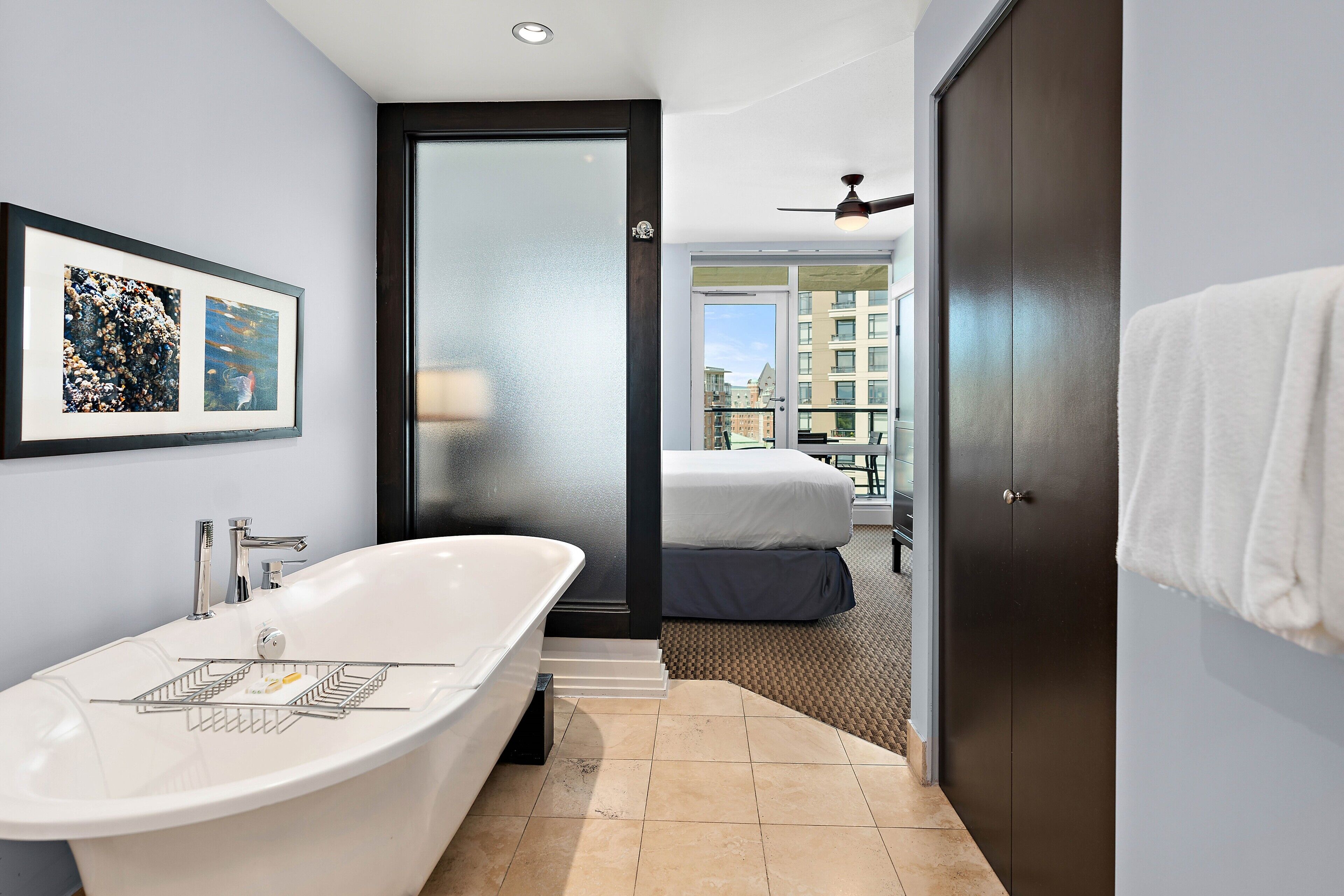 Une salle de bain moderne avec une baignoire sur pieds, une œuvre d’art encadrée au mur et une vue sur la ville par la porte.