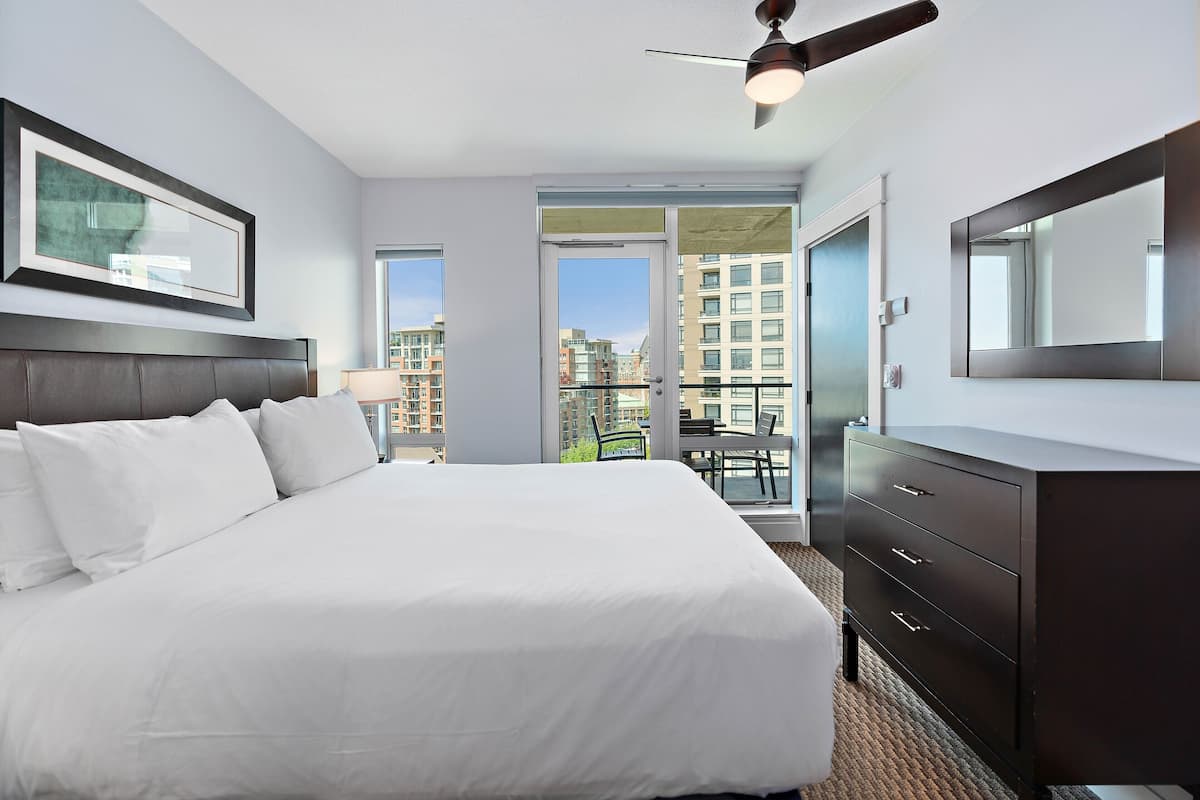 A hotel room with a large bed, a dark dresser, a ceiling fan, and a view of the city through a large window.