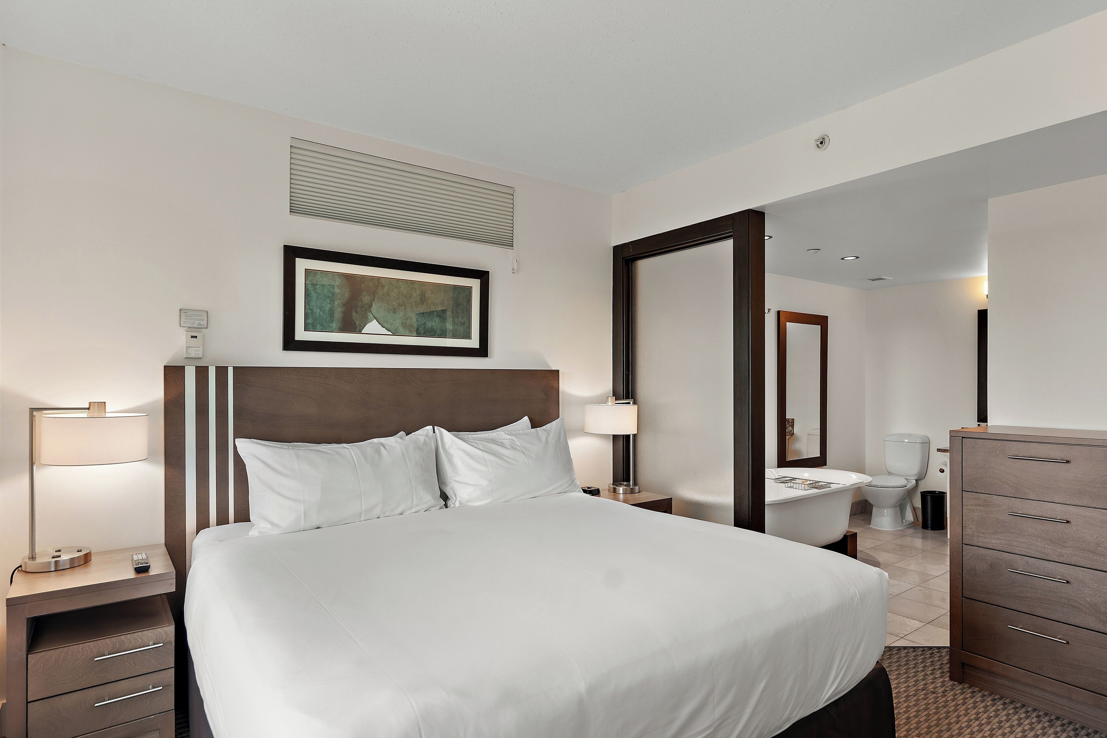 Une chambre d’hôtel avec un grand lit, des tables de chevet, une petite table, une lampe, un tableau encadré au mur, une salle de bain avec des toilettes et un lavabo, et une commode en bois.